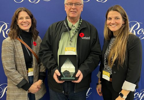 Three people standing in front of a backdrop with the word 'Royal' printed several times across it. The middle person his holding an award.