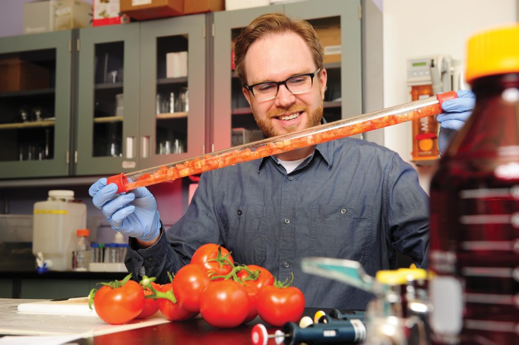 A man with glasses and a beard holding a clear tube with chopped tomatoes inside. In front of him are some whole tomatoes on the vine.